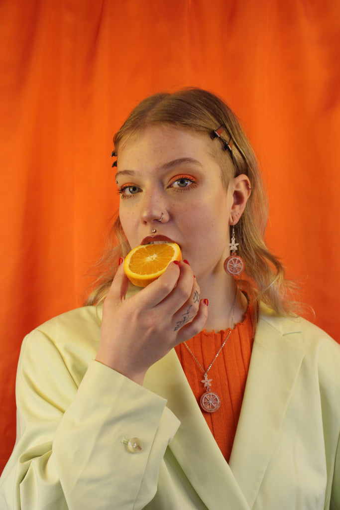 model wearing orange slice pendant suspended from white flower charms on stainless steel earwires, eating an orange