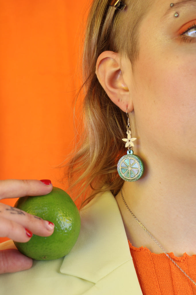 Model wearing Glittery lime fruit slice charm suspended from a white flower on stainless steel earwires