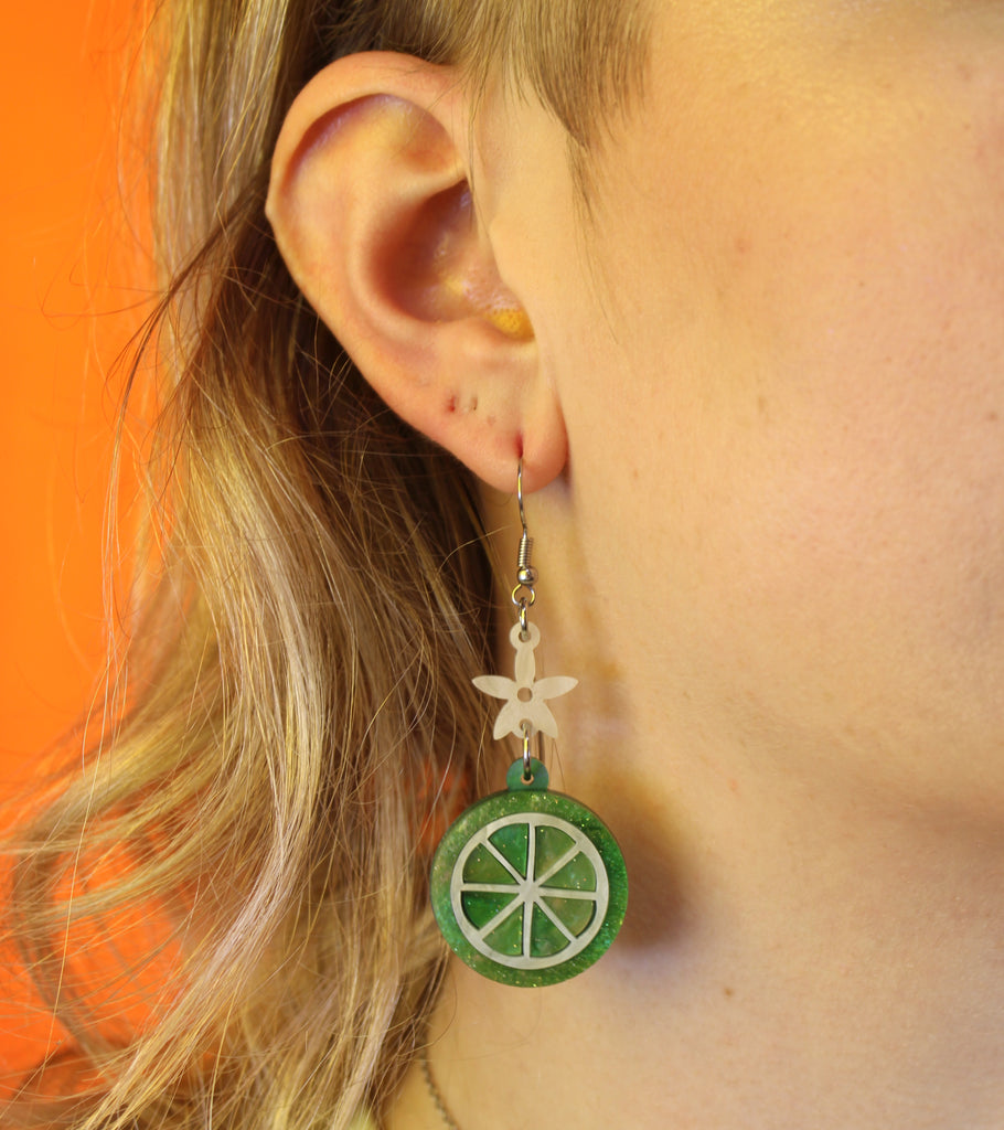 Model wearing Glittery lime fruit slice charm suspended from a white flower on stainless steel earwires