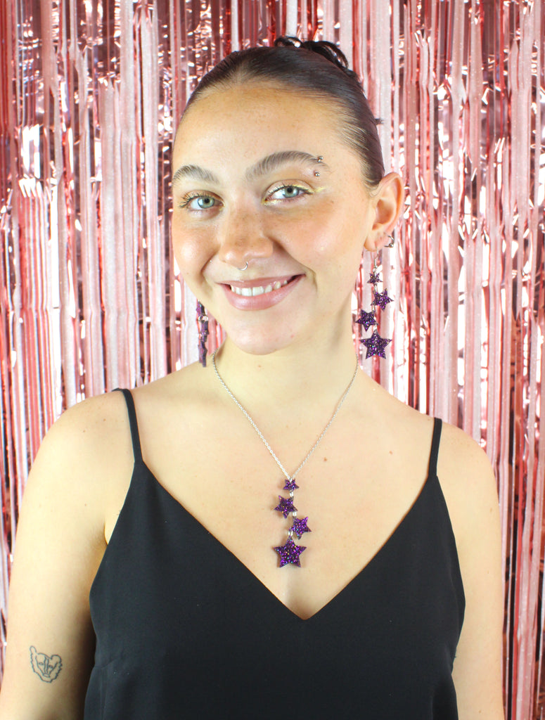 Model wearing Four purple glitter star charms with gold fleck detailing hanging from stainless steel earwires, varying in size (the largest one hanging at the bottom) Model is also wearing the matching necklace