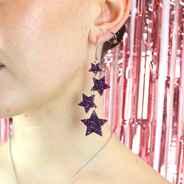 Model wearing Four purple glitter star charms with gold fleck detailing hanging from stainless steel earwires, varying in size (the largest one hanging at the bottom) Model is also wearing the matching necklace