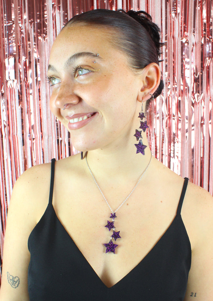 Model wearing Four purple glitter star charms with gold fleck detailing hanging from stainless steel earwires, varying in size (the largest one hanging at the bottom) Model is also wearing the matching necklace