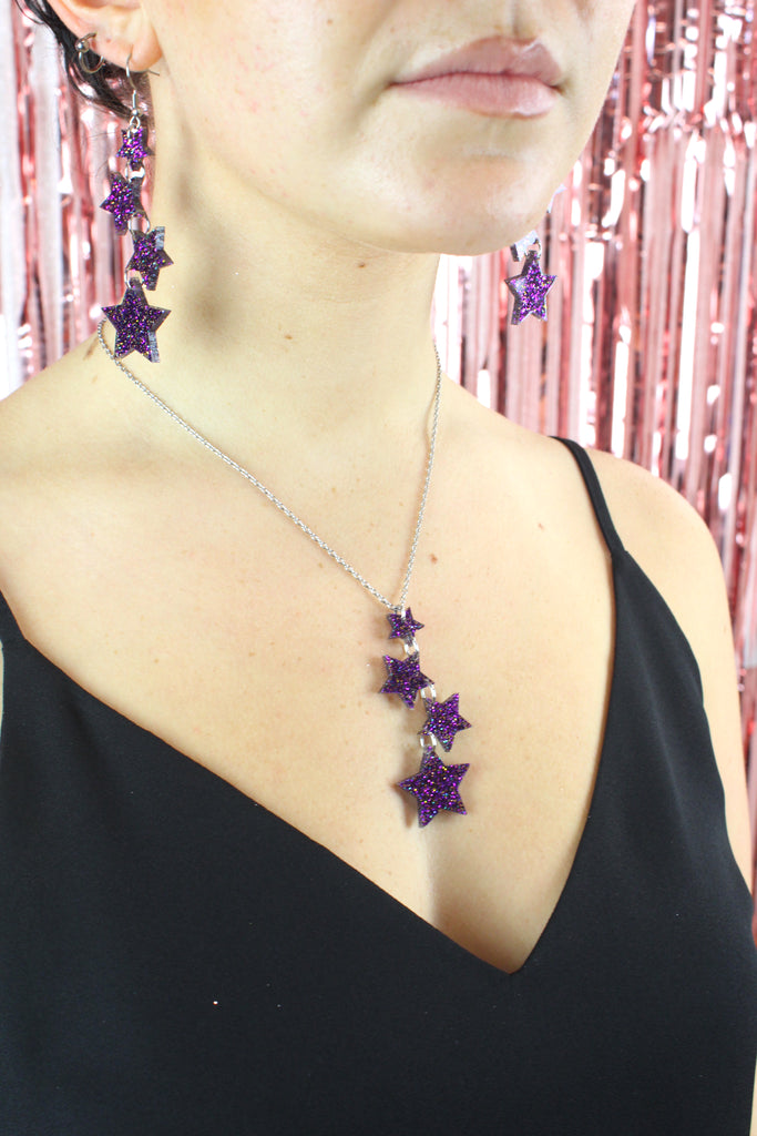 Model wearing Four purple glitter star charms with gold fleck detailing hanging from stainless steel earwires, varying in size (the largest one hanging at the bottom). Model is also wearing the matching necklace